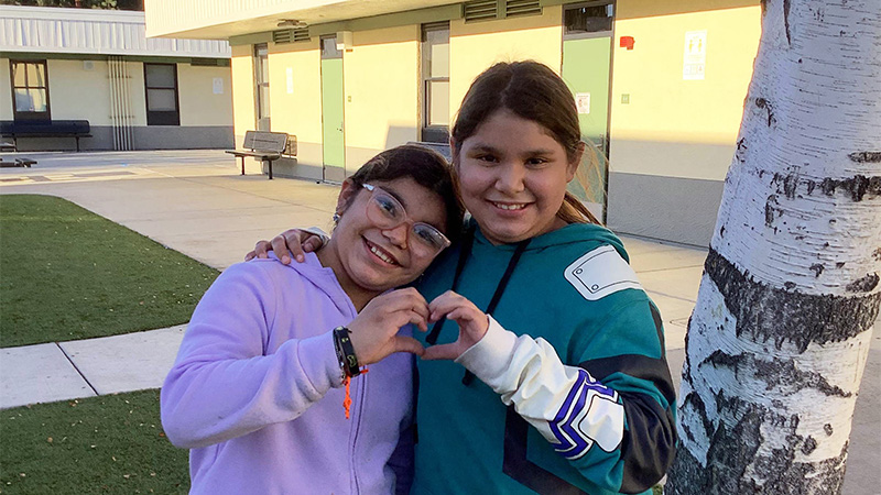 Outdoor image of sisters with heart hands in front of a school building